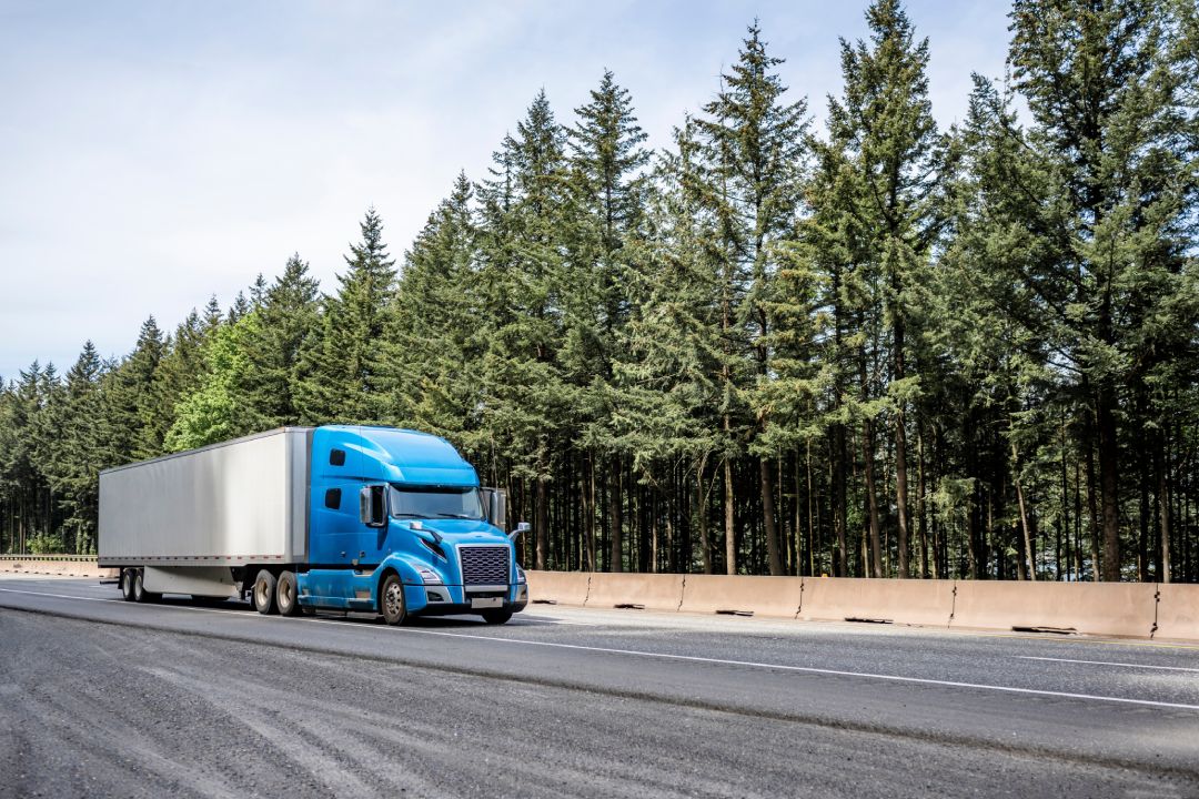 Blue big rig long haul industrial semi truck tractor transporting cargo in dry van semi trailer running on curving highway road with protective fence and green mountain forest in Columbia Gorge