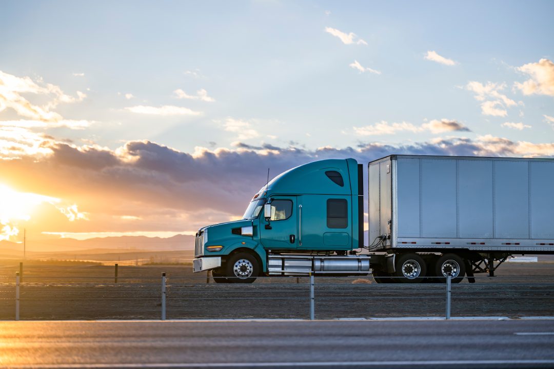 Industrial de transporte de gran camión verde semirremolque tractor con piezas de cromo el transporte de carga comercial en cargado semirremolque furgoneta seca que se ejecuta en el camino de la carretera en el momento de la puesta del sol en California