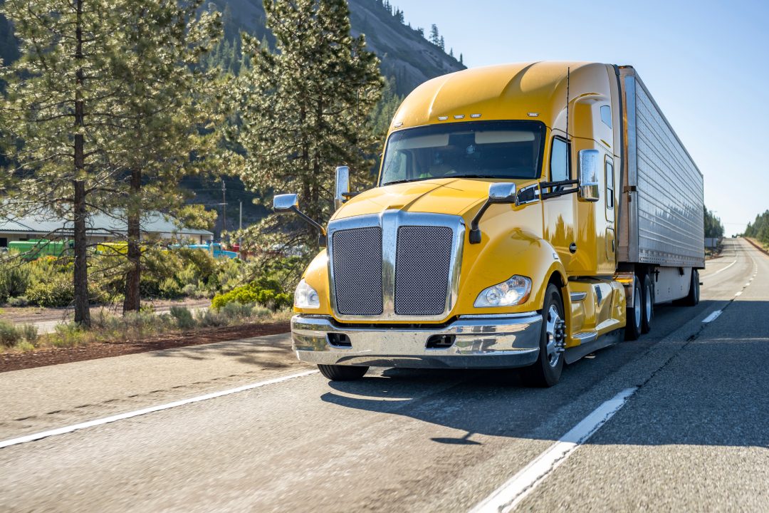 Classic long haul big rig yellow semi truck tractor with truck driver cab sleeping compartment transporting cargo in refrigerator semi trailer running on the highway road with rest area on the side