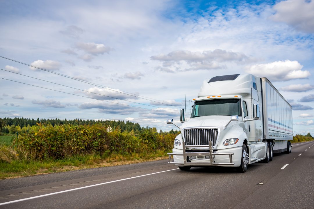 Potente gran plataforma de grado industrial blanco de alta cabina diesel semi camión con parrilla de tuberías de guardia de transporte de carga comercial en seco semirremolque furgoneta que se ejecuta en el camino de la carretera con la colina verde