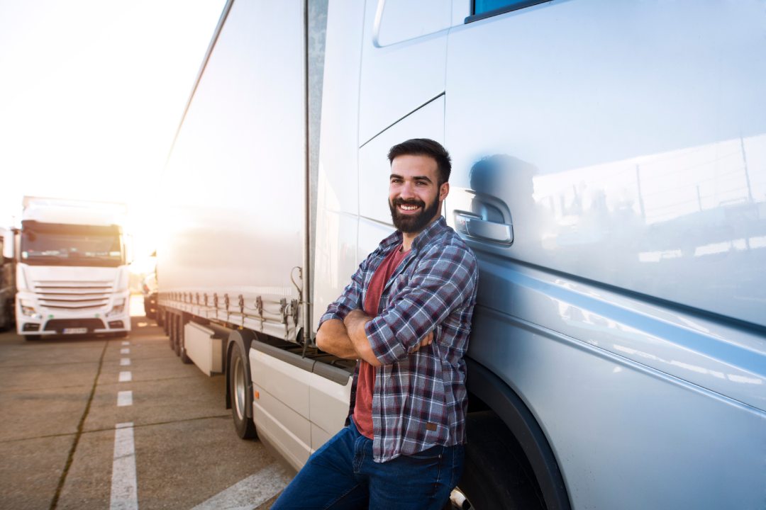Portrait of young Caucasian bearded trucker with arms crossed standing by his truck vehicle. Transportation service. Truck driver job.